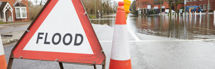 Image of flood warning sign and cones on a flooded road, the road is running through a town in the UK